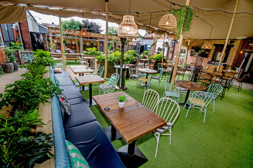 Covered outdoor courtyard at The Hart in Headless Cross, Redditch, featuring blue cushioned bench seating, wooden dining tables, wicker pendant lights and raised green planters.