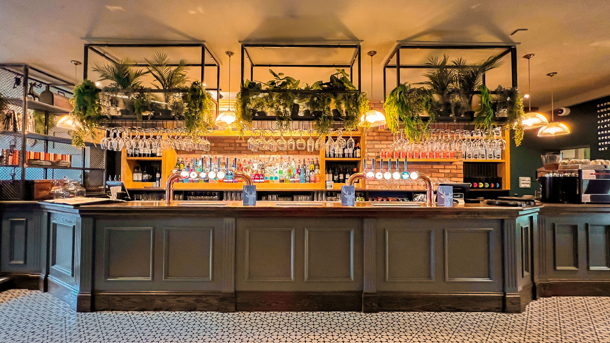 The Hart pub bar in Headless Cross, Redditch, featuring a dark green traditional bar counter, exposed brickwork, copper beer taps, hanging glassware and decorative greenery.