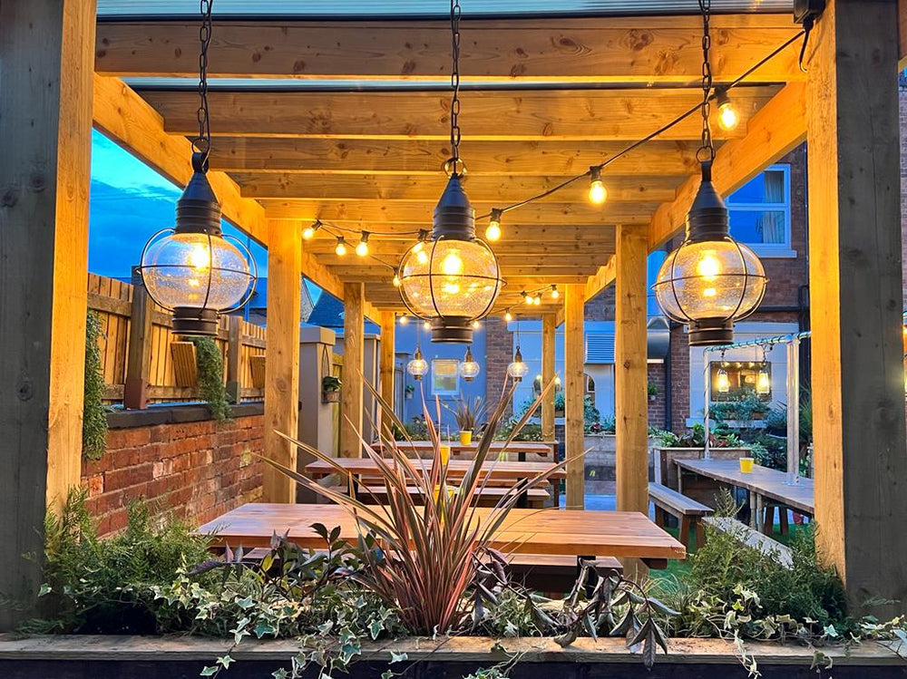 Sheltered beer garden seating at The Hart Pub in Redditch, with timber beams, hanging lanterns, wooden picnic tables and leafy borders.